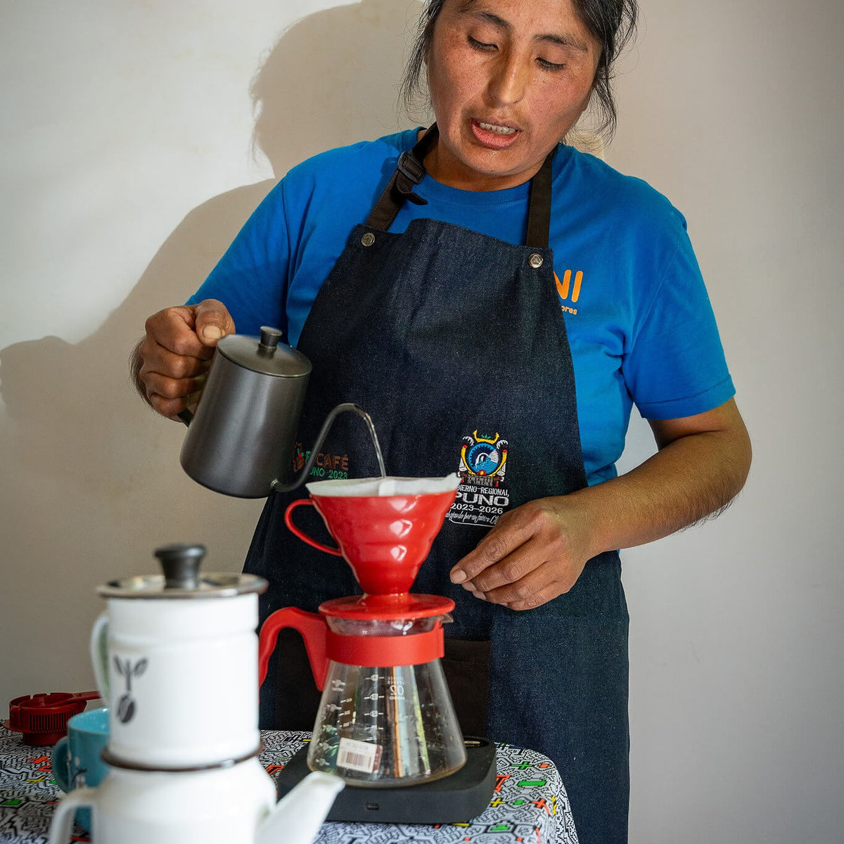 Person in a blue shirt and apron preparing coffee using a pour-over setup.